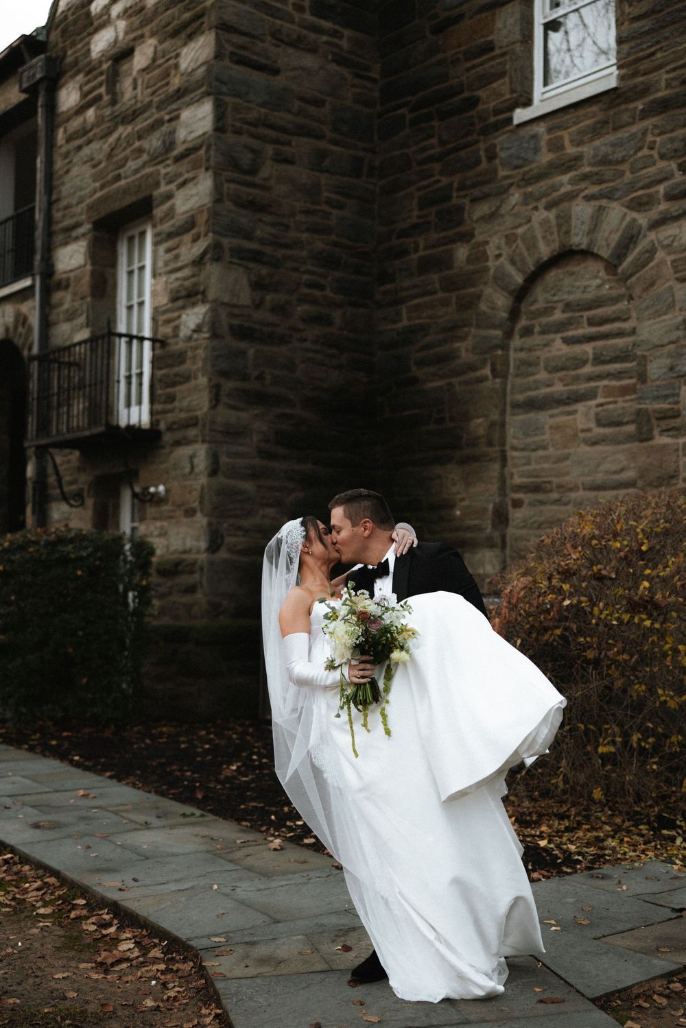 Groom lifts bride for a kiss outside a historic stone building on their wedding day.