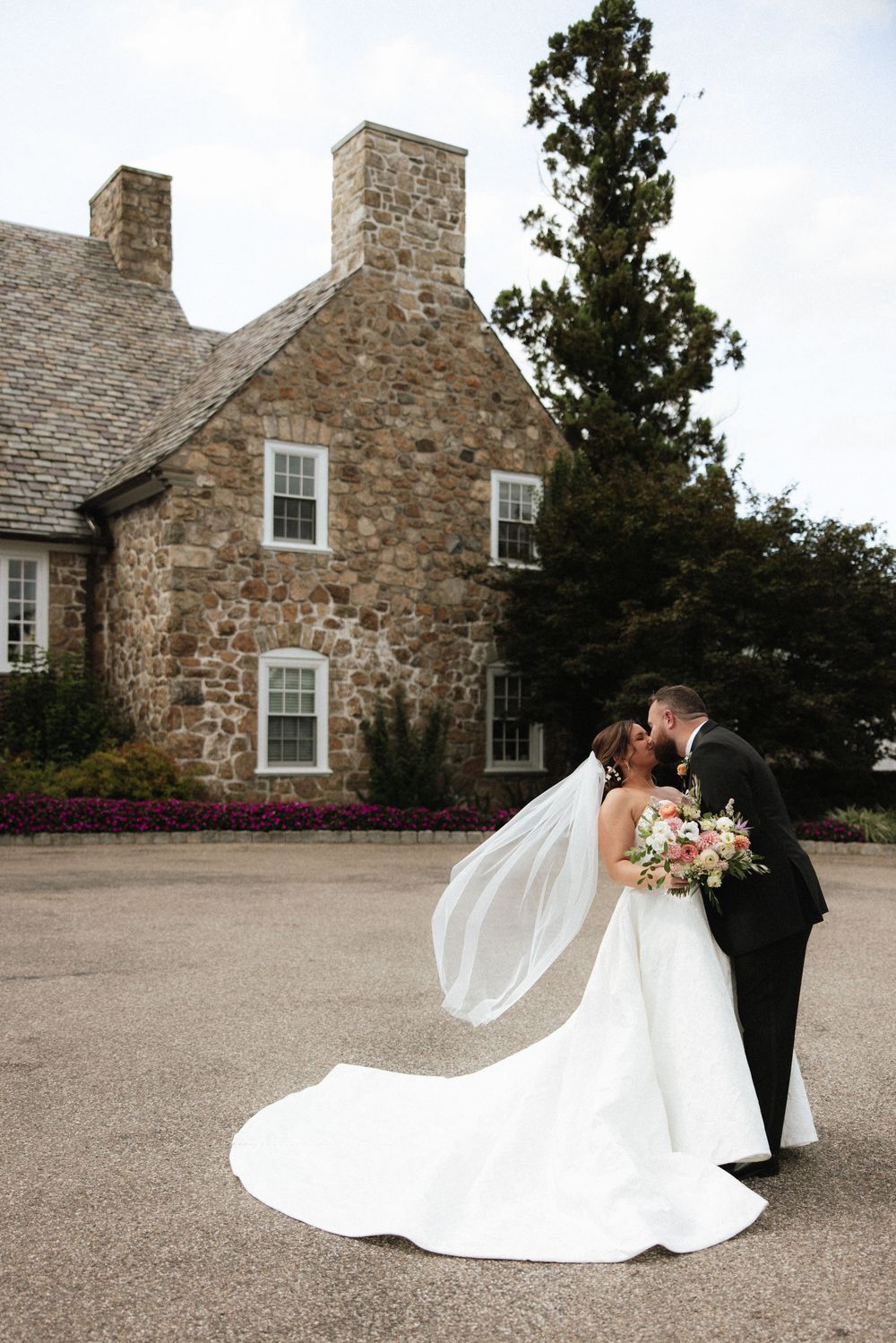 Bride and groom share a kiss outside a stone mansion, with the bride's veil flowing in the wind.