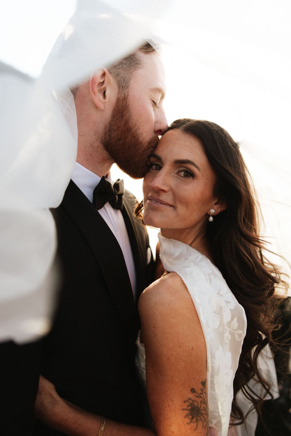 Groom kisses bride on the cheek as her veil blows in the wind during outdoor wedding portraits.