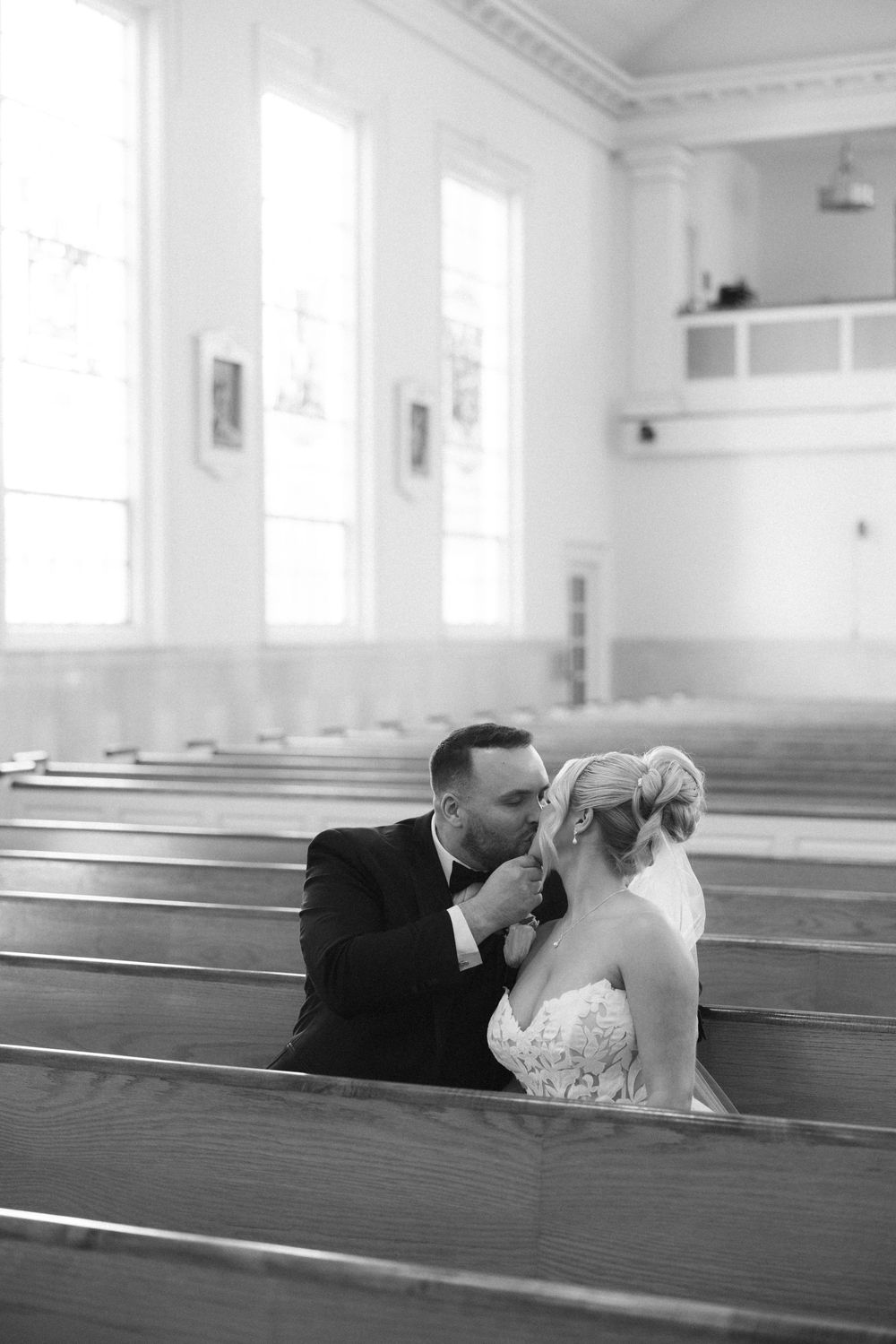 Black and white photo of groom kissing bride in church pew with bright windows in background.