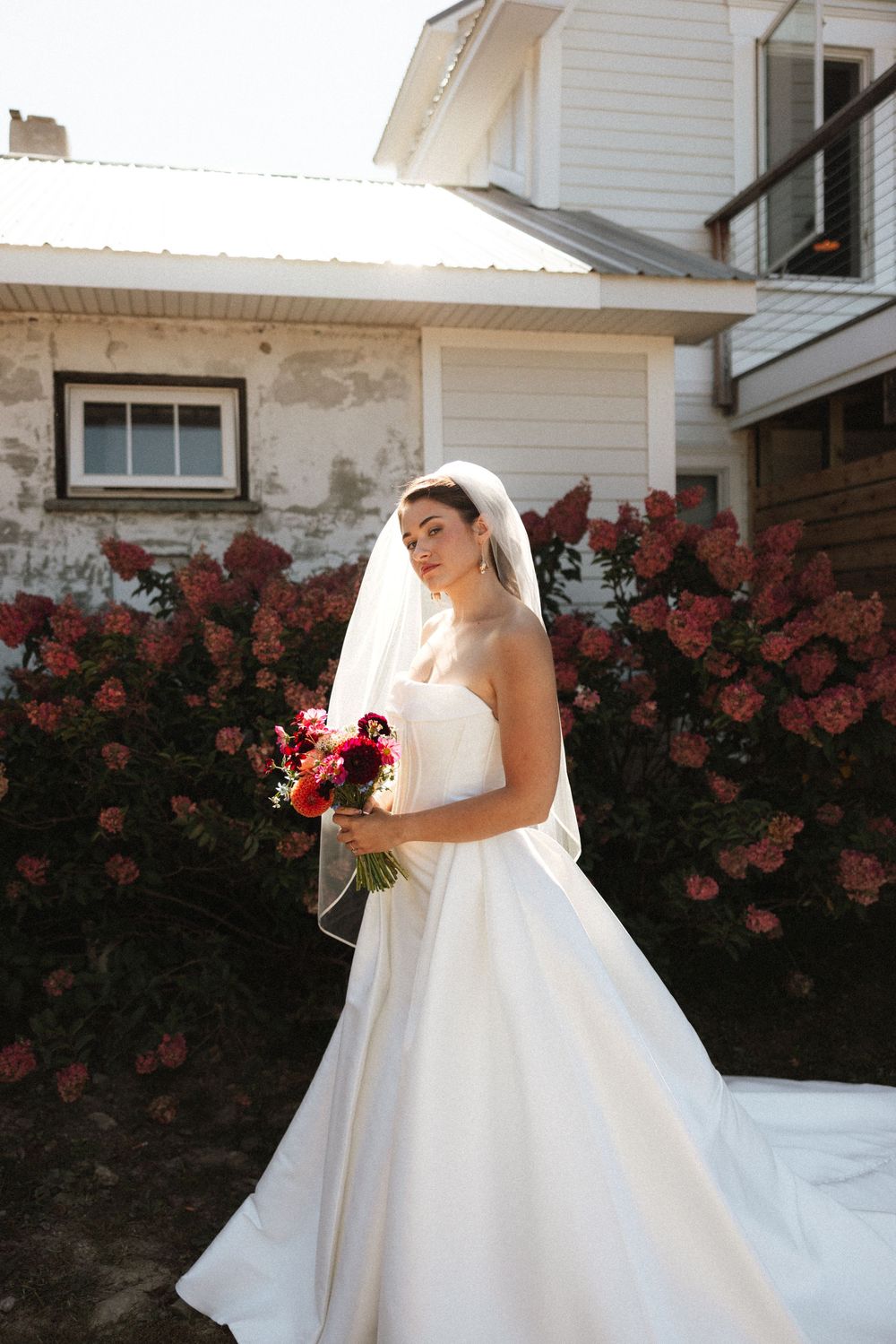 Bride in strapless white gown holds bouquet in front of blooming pink hydrangeas at sunset.