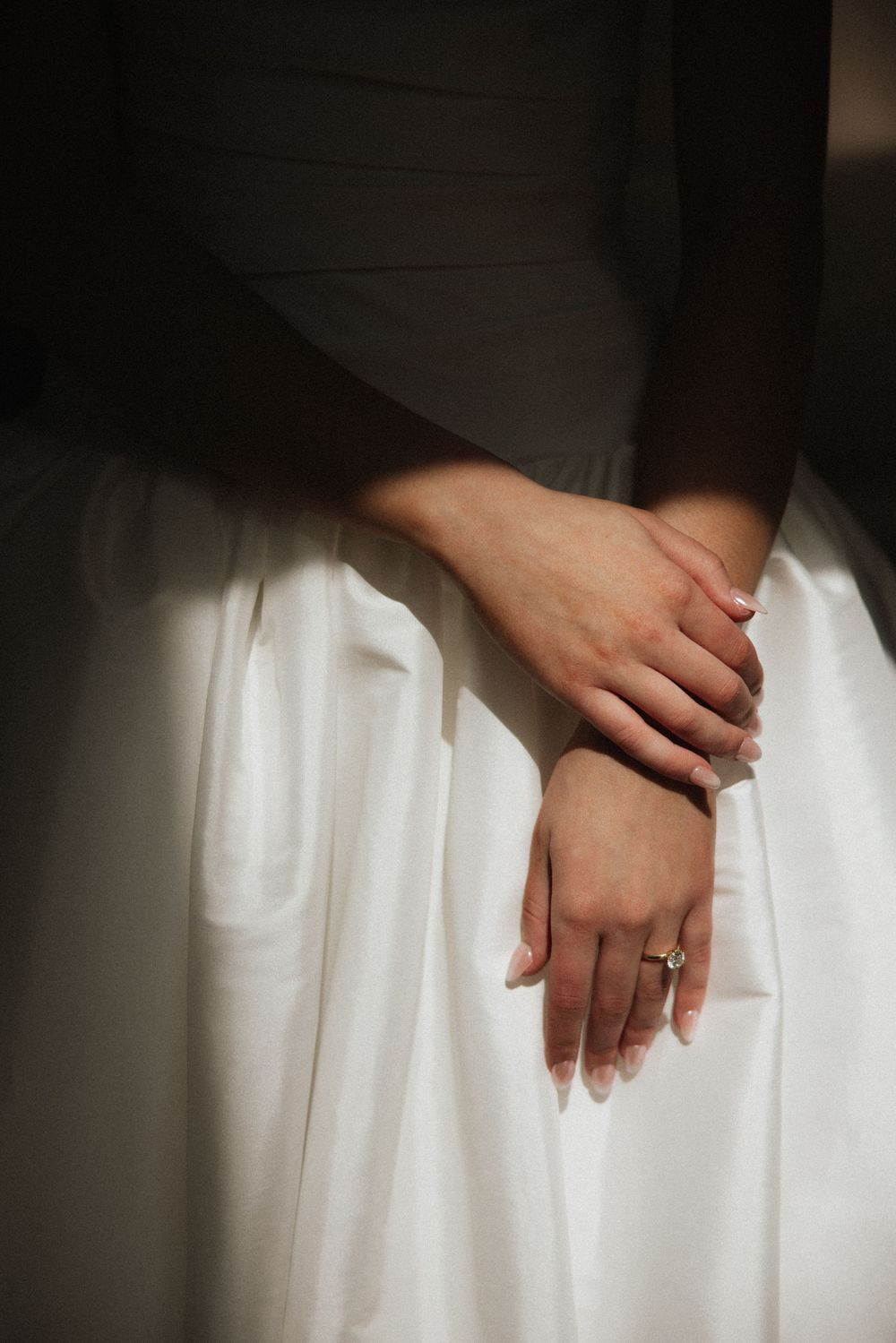 Close-up of bride's hands clasped over white wedding gown with diamond engagement ring in sunlight.
