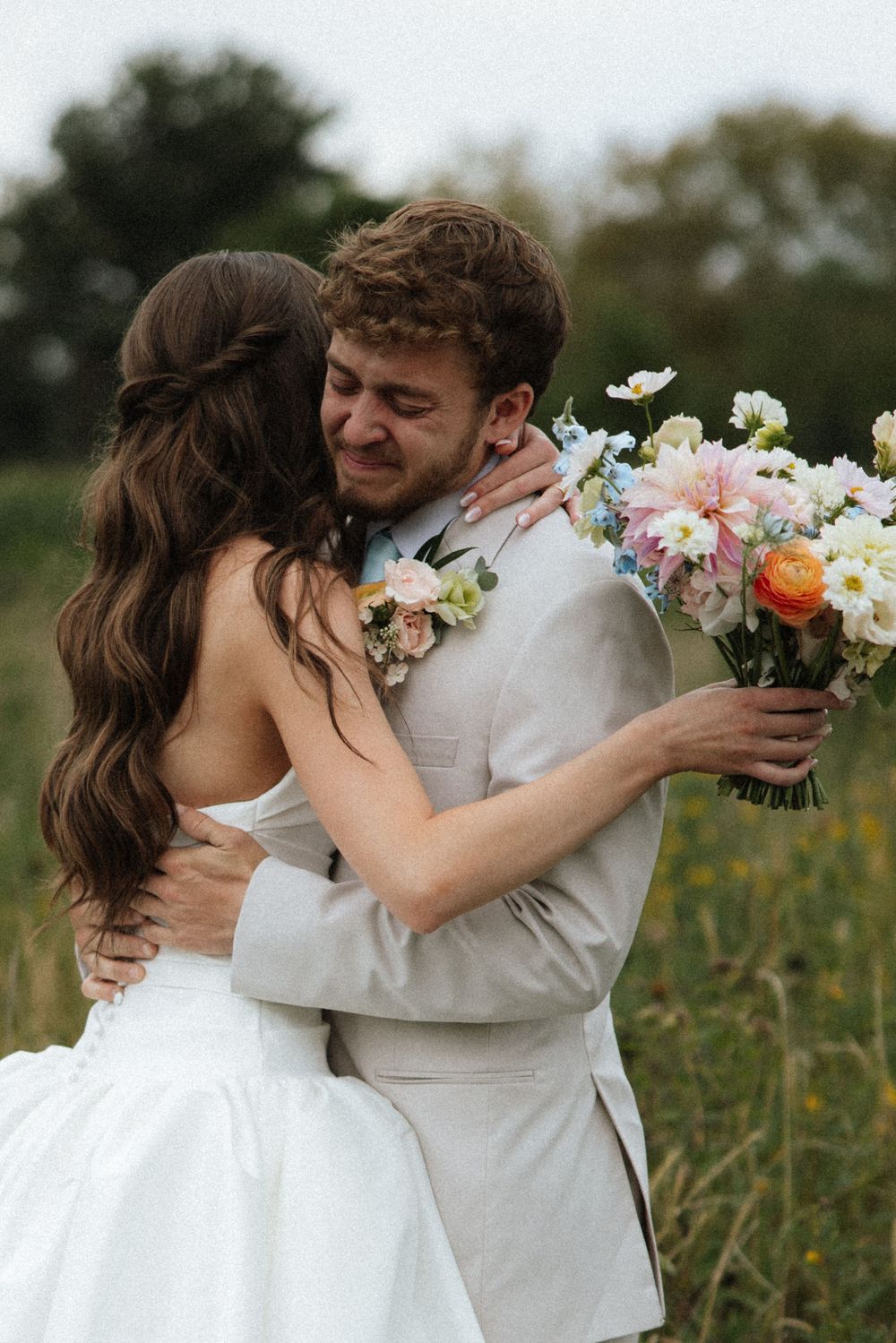 Emotional groom in beige suit hugging bride in white dress while holding colorful bouquet in field.