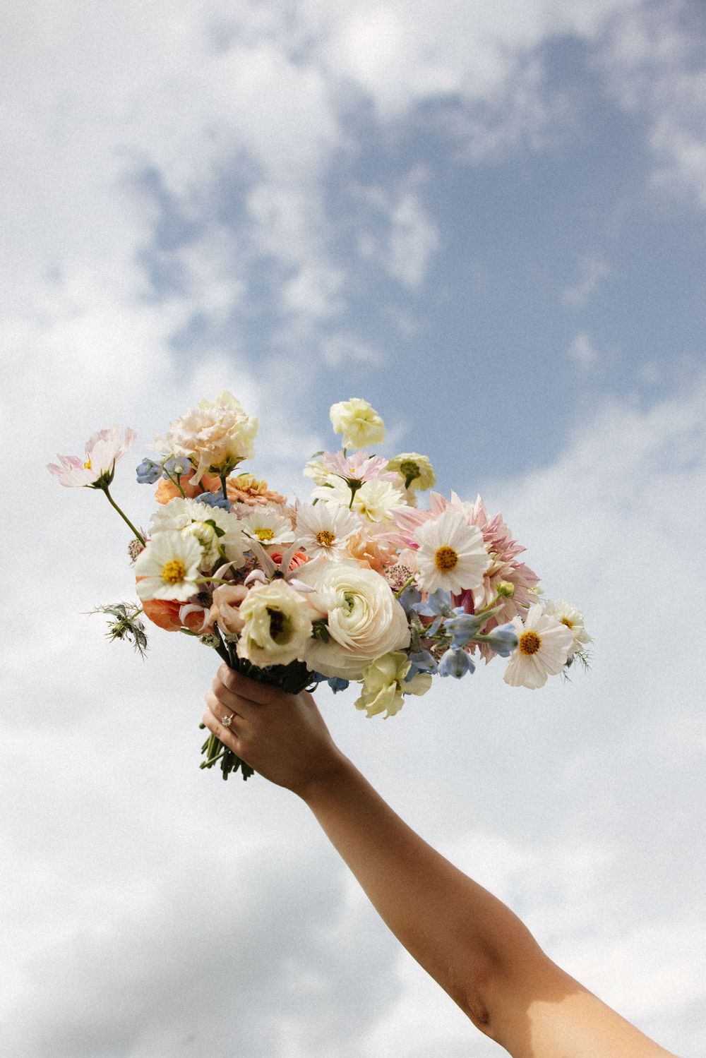Hand holding a colorful bridal bouquet of daisies, ranunculus, and wildflowers against cloudy sky.