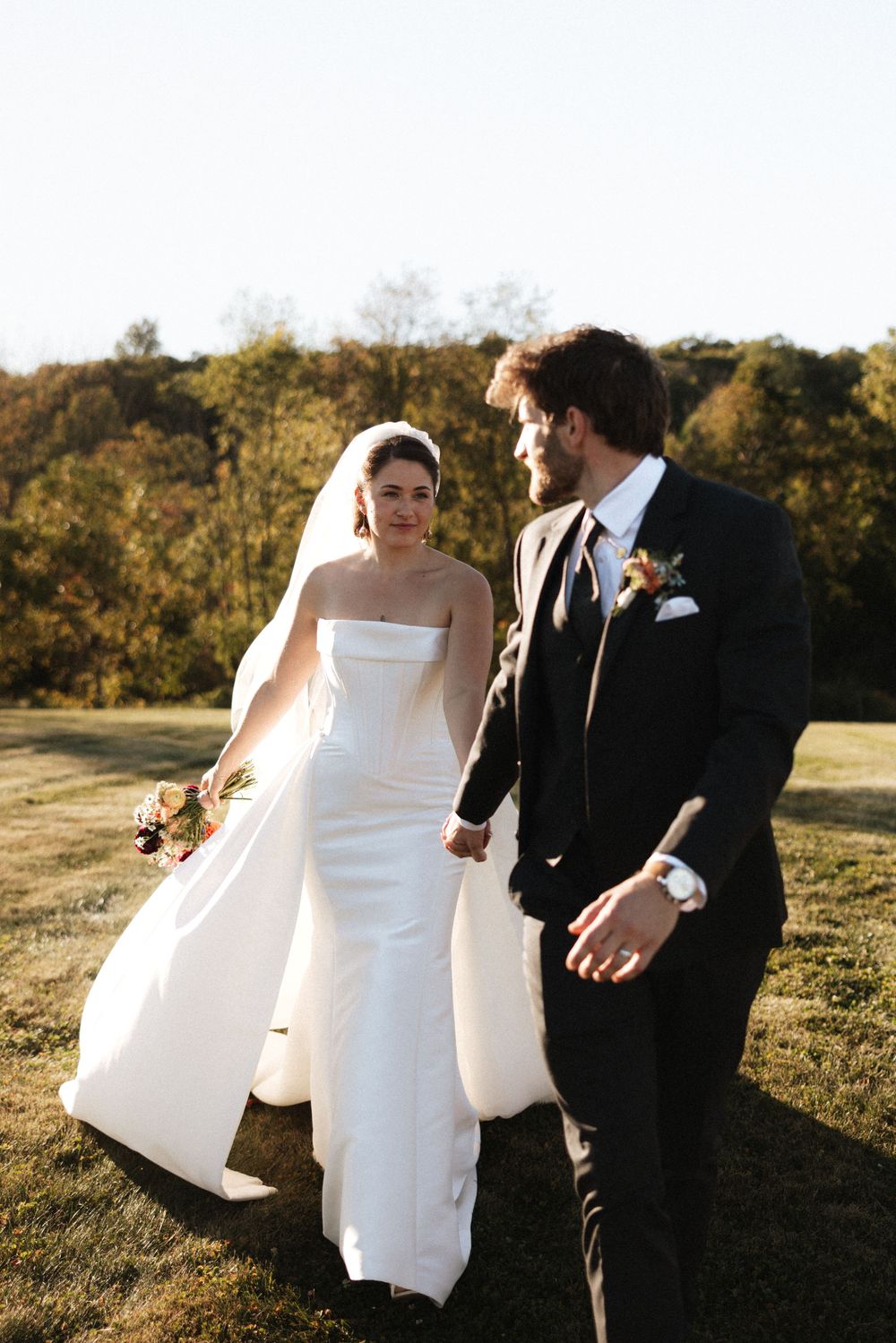 Bride in white strapless gown and groom in black suit walk hand in hand across sunlit green lawn.