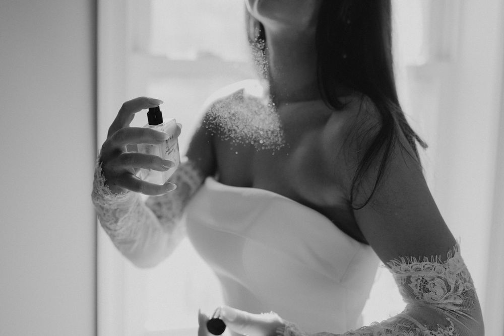 Black and white photo of tattooed bride in strapless gown applying perfume spray on her wedding day.