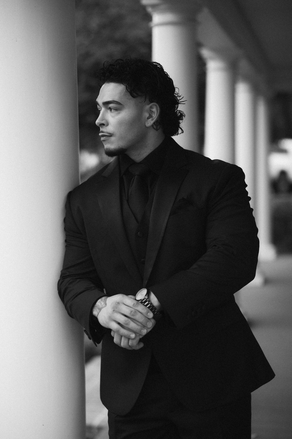 Black and white portrait of groom in black suit leaning against white column looking away thoughtfully.
