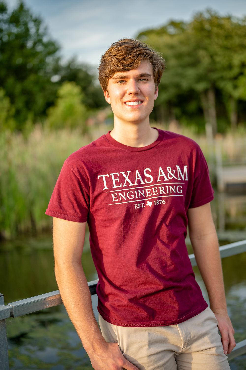 Senior Student wearing Texas A&M Engineering t-shirt stands outdoors near water.