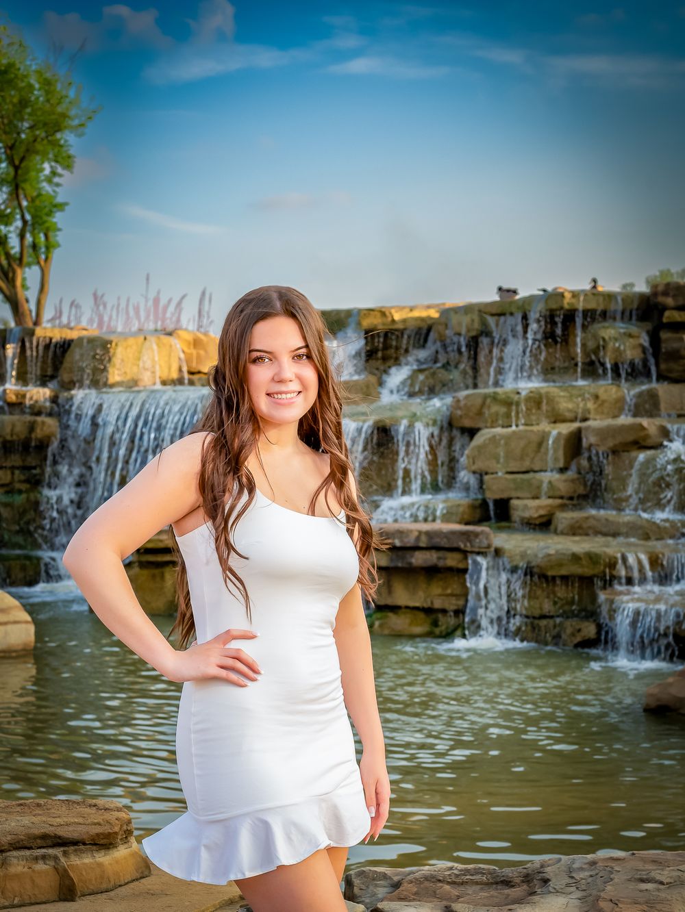 High school senior wearing white summer dress portrait by cascading stone waterfall feature under blue sky.