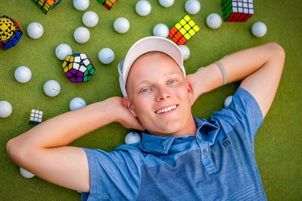 Teen lying on golf green surface surrounded by white balls and colorful Rubik's cubes.