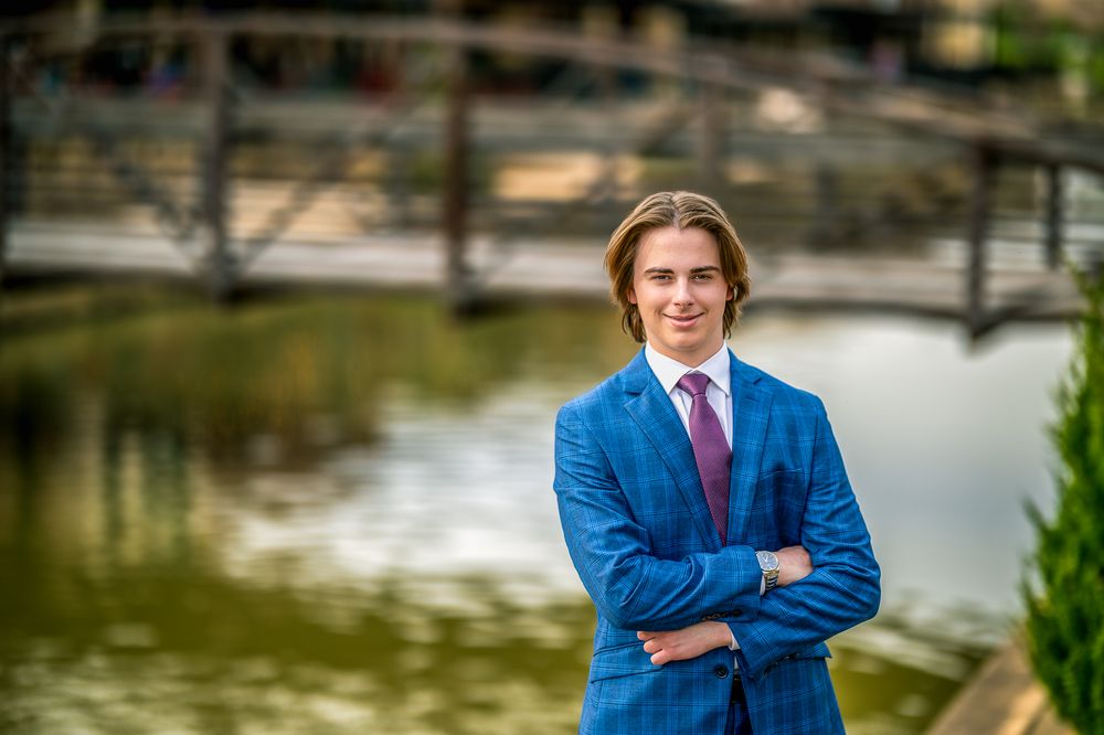 Senior Portrait in blue suit standing by waterfront with decorative bridge in background.