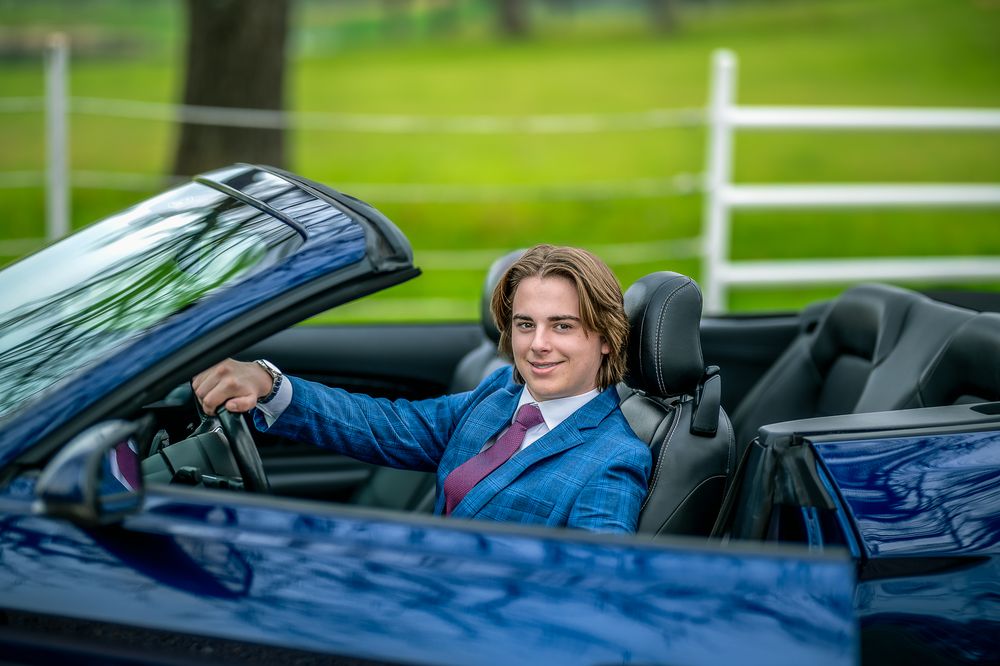 Senior portrait in Blue convertible sports car parked on grass with white fence in background.