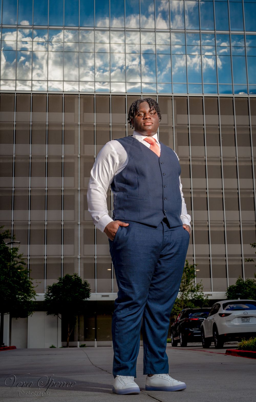 Professional senior portrait showcasing business attire against modern glass building and cloudy sky.