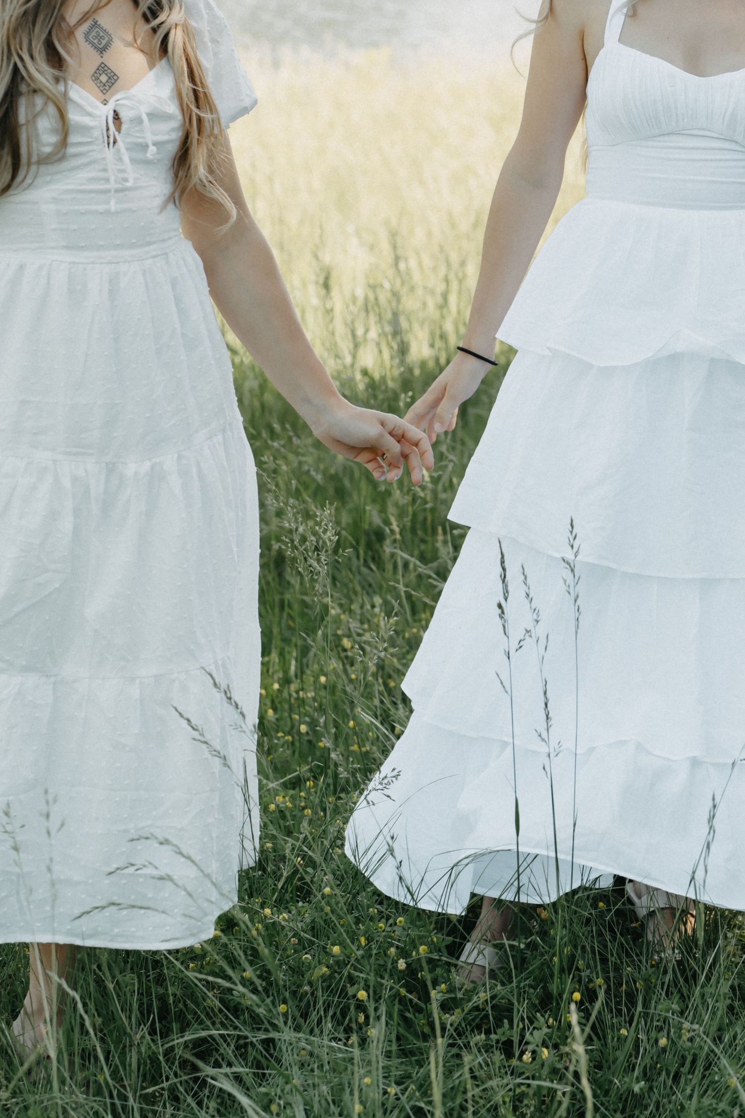 Two women in white dresses holding hands while walking through a lush green meadow.
