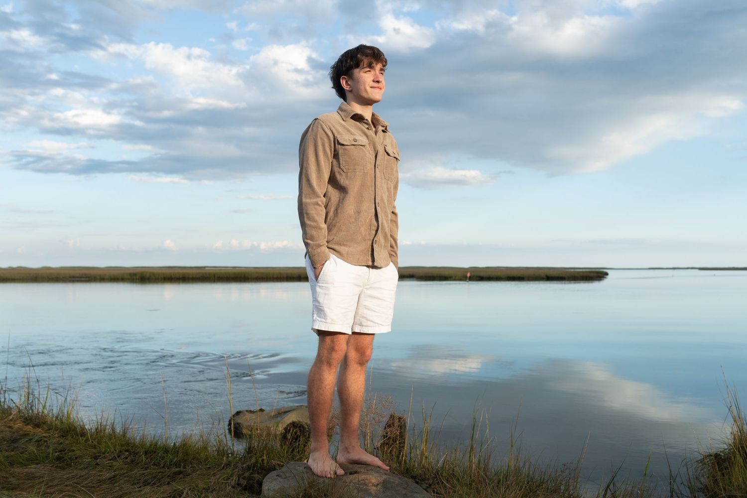 Person in tan jacket standing near water's edge with cloudy sky and calm lake in background.