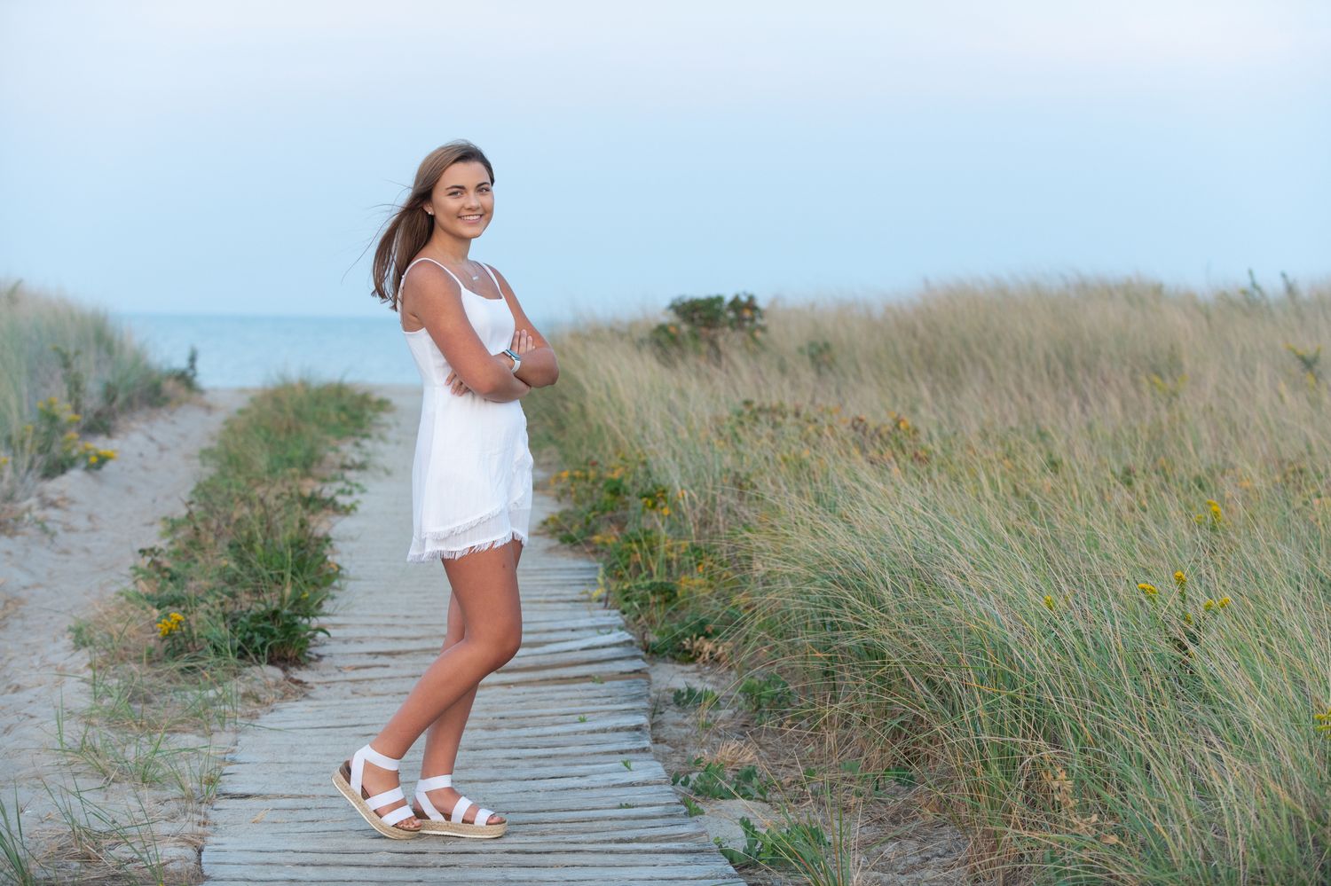 Woman in white dress walking along wooden boardwalk through beach grass at sunset.