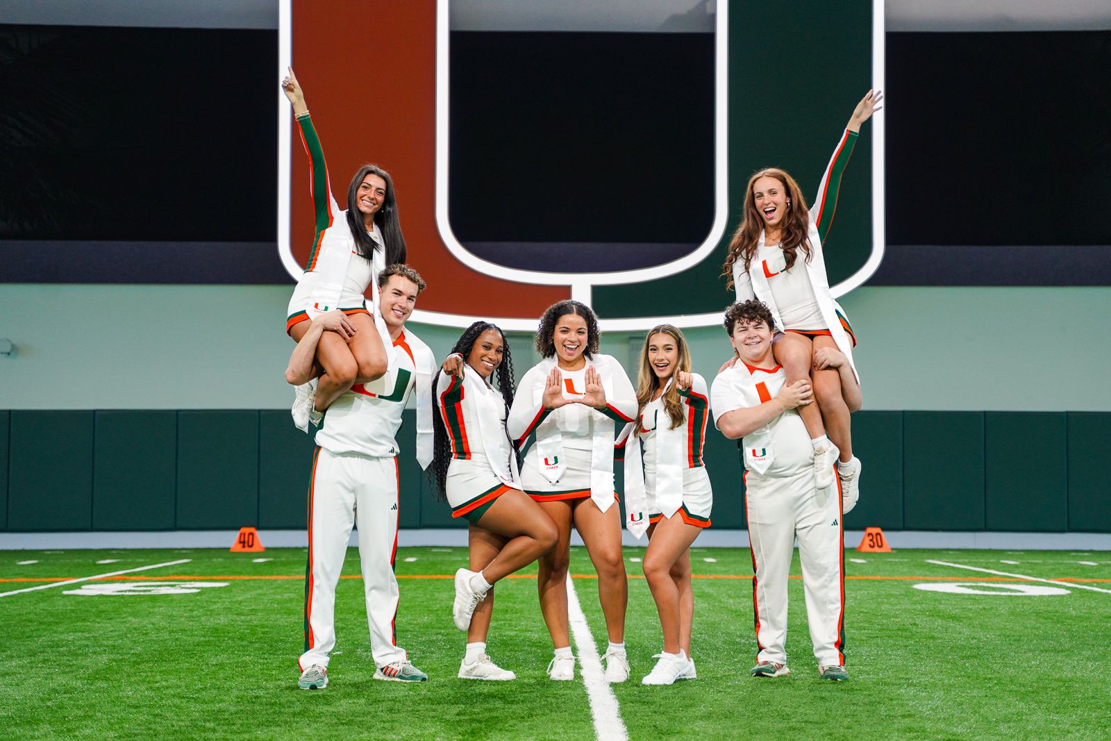Cheerleaders perform stunts on a football field with University of Miami logo.