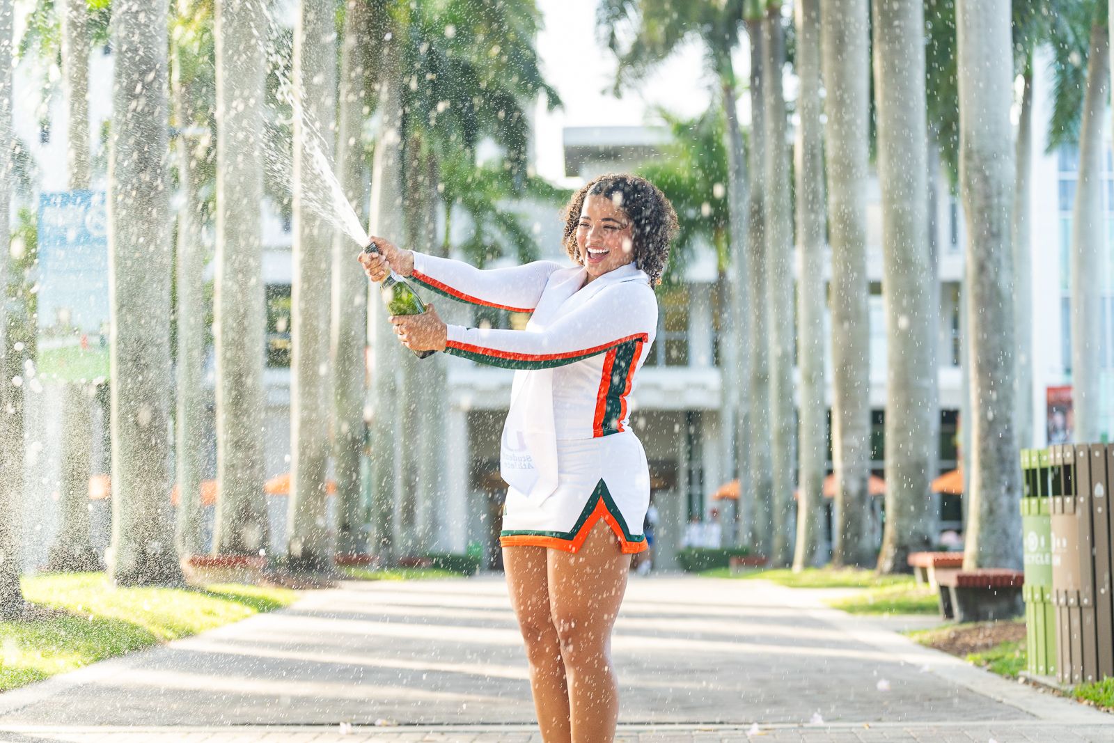 Graduate celebrating with champagne spray in front of palm tree-lined walkway at University of Miami.