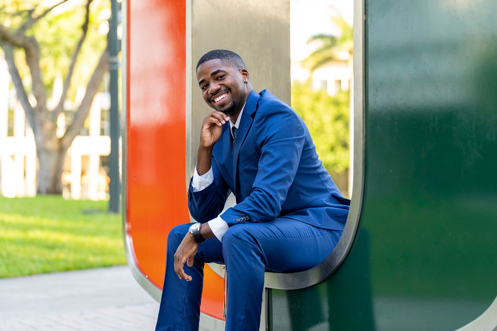 Professional in blue suit sits thoughtfully by University of Miami logo outdoors.