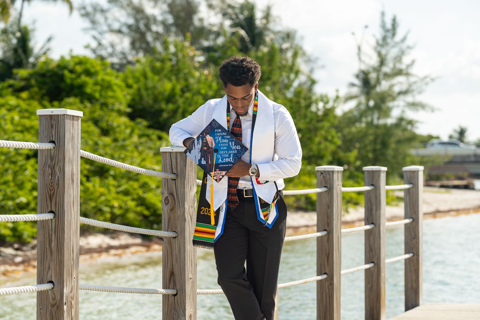 Graduate in white coat poses with stethoscope on wooden boardwalk by the ocean.