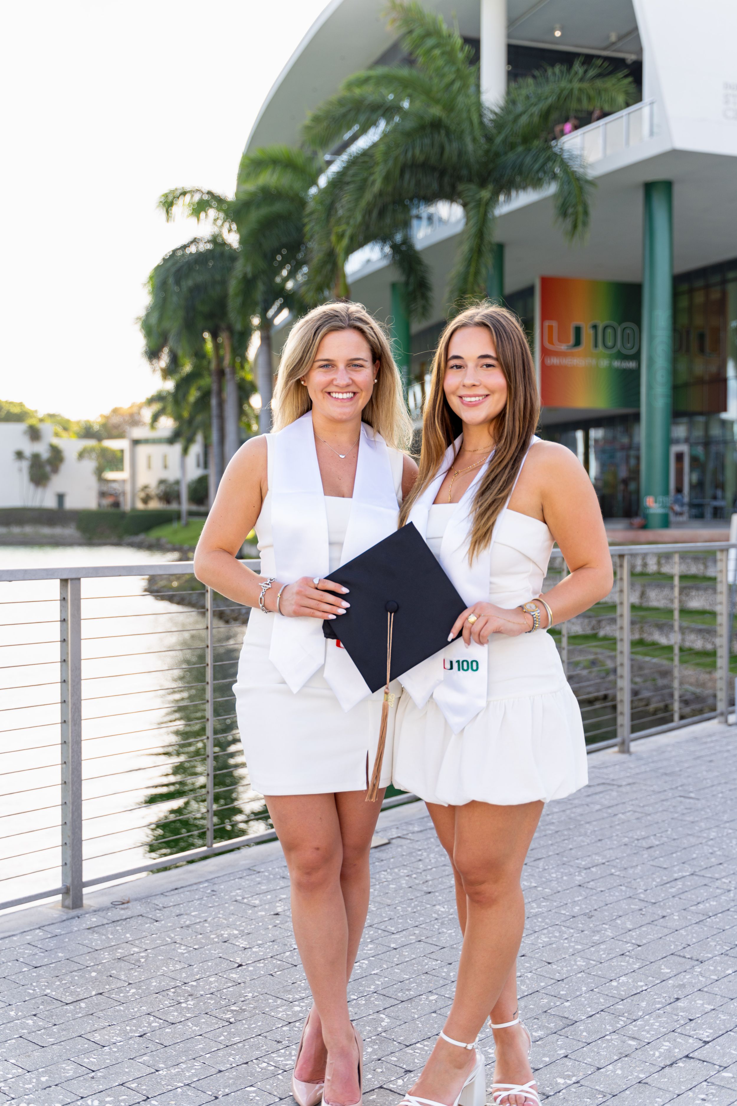 Two friends in summer outfits posing together on a walkway with palm trees and buildings in the background.