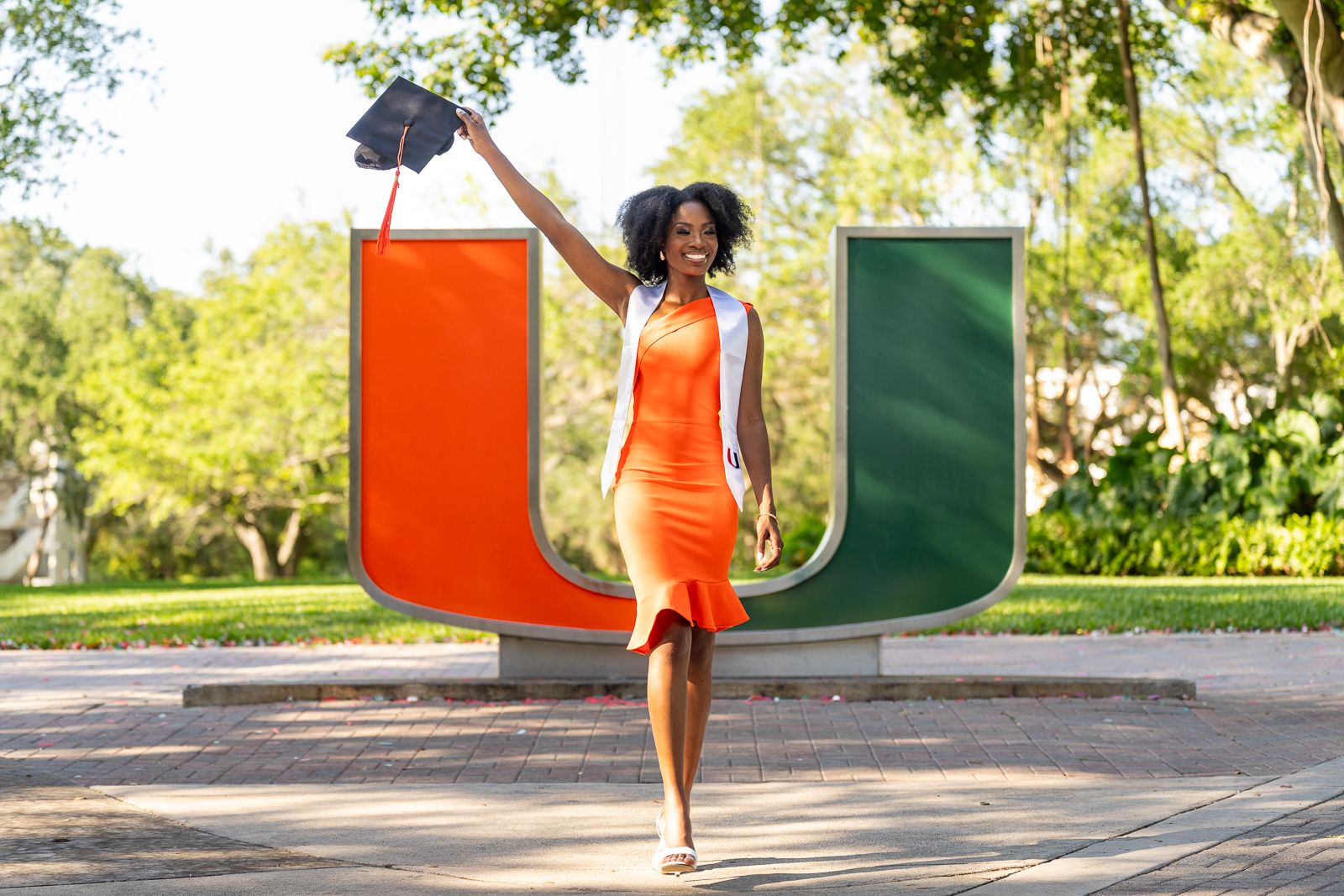 Graduate in orange dress celebrates with cap by University of Miami logo on sunny day.