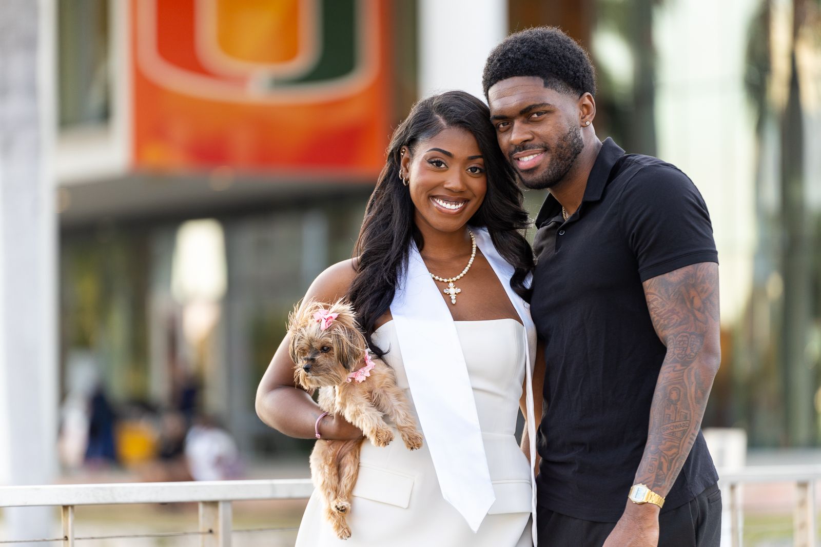 Couple poses with small dog outside campus building with University of Miami banner visible.