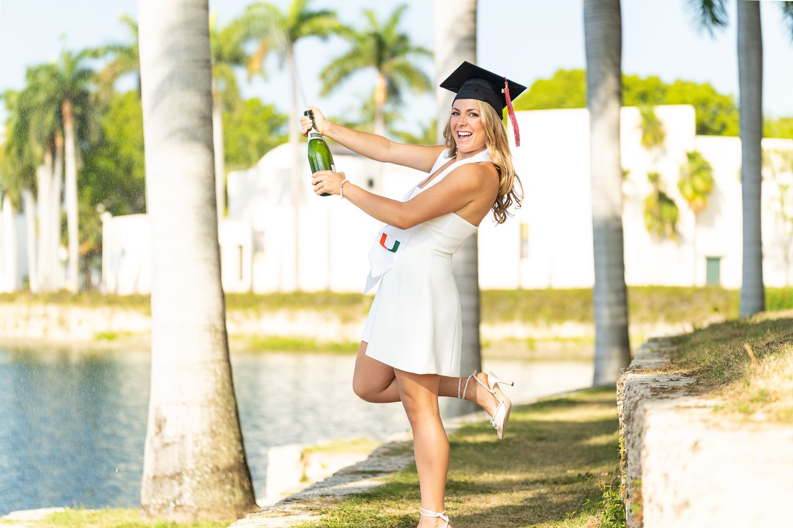 Graduate in white dress and cap celebrating with champagne bottle by palm trees and lake.