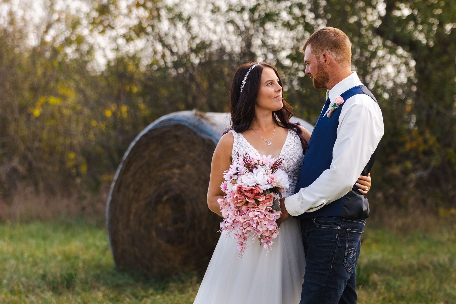 Wedding couple embracing in countryside setting with hay bale backdrop.