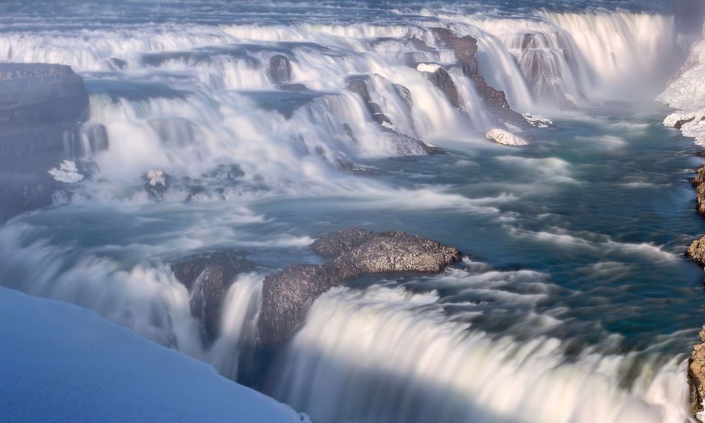 Aerial view of massive waterfalls flowing over icy cliffs with mist rising from turbulent waters.