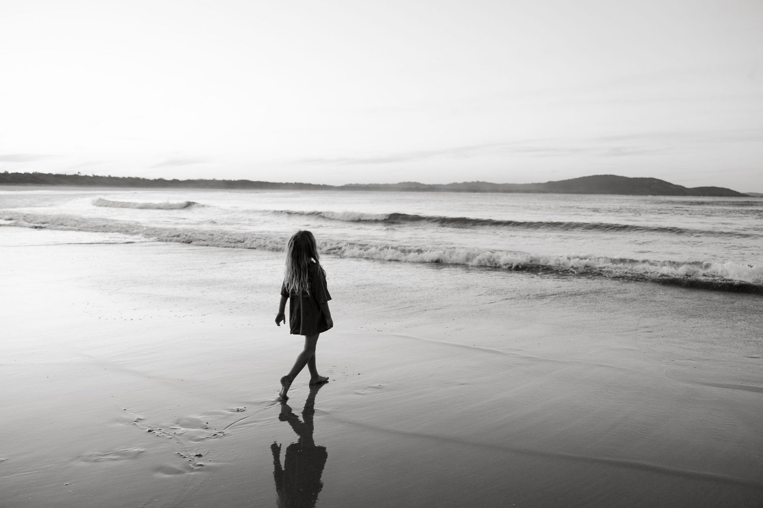 Lone figure walking on empty beach creating reflection in wet sand during overcast day.