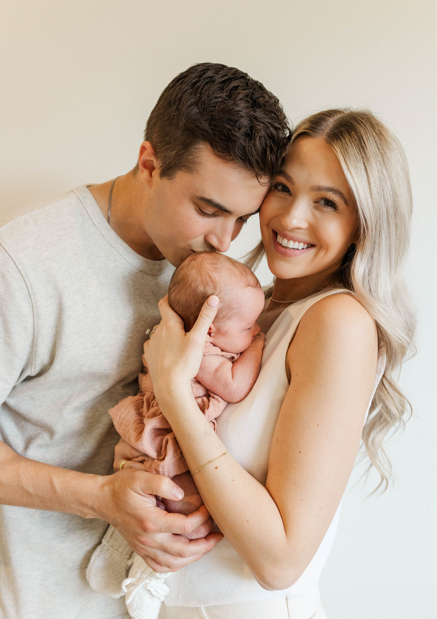 Couple smiling and kissing newborn baby in soft neutral tones indoors.