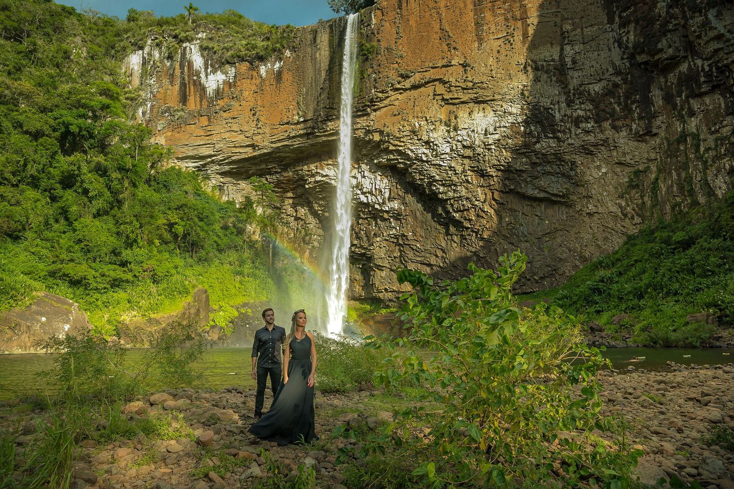 Two silhouettes stand against dramatic waterfall backdrop in natural setting.