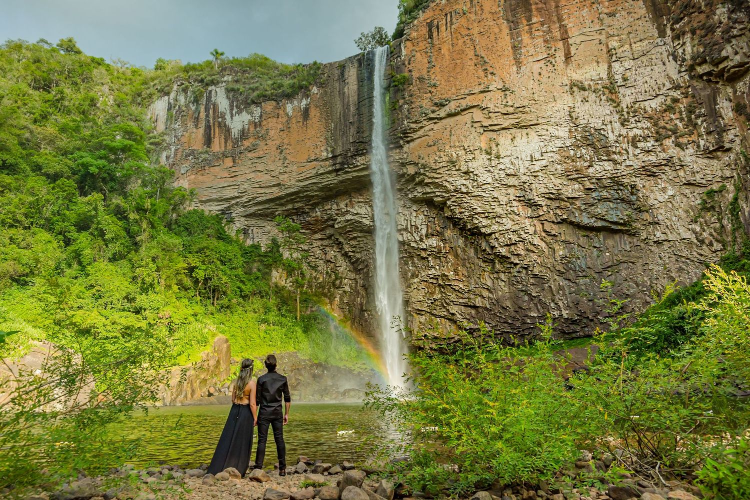 Two figures admire a tall waterfall cascading down rocky cliffs with rainbow visible.