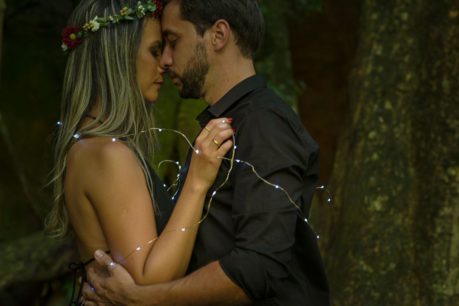 A romantic couple embraces in a dimly lit forest setting with fairy lights creating a magical atmosphere.
