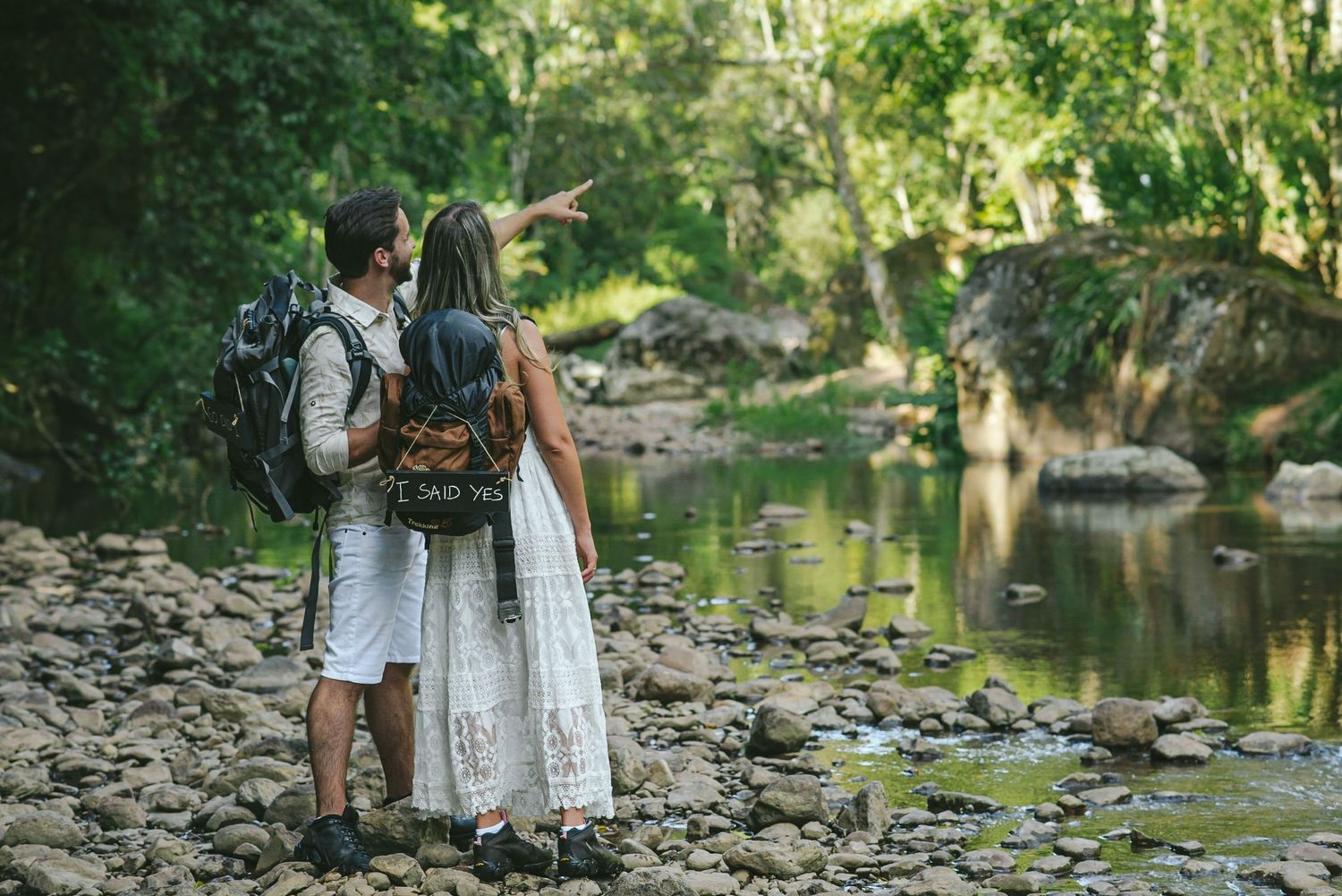 Couple with backpacks standing by a rocky stream in a green forest.