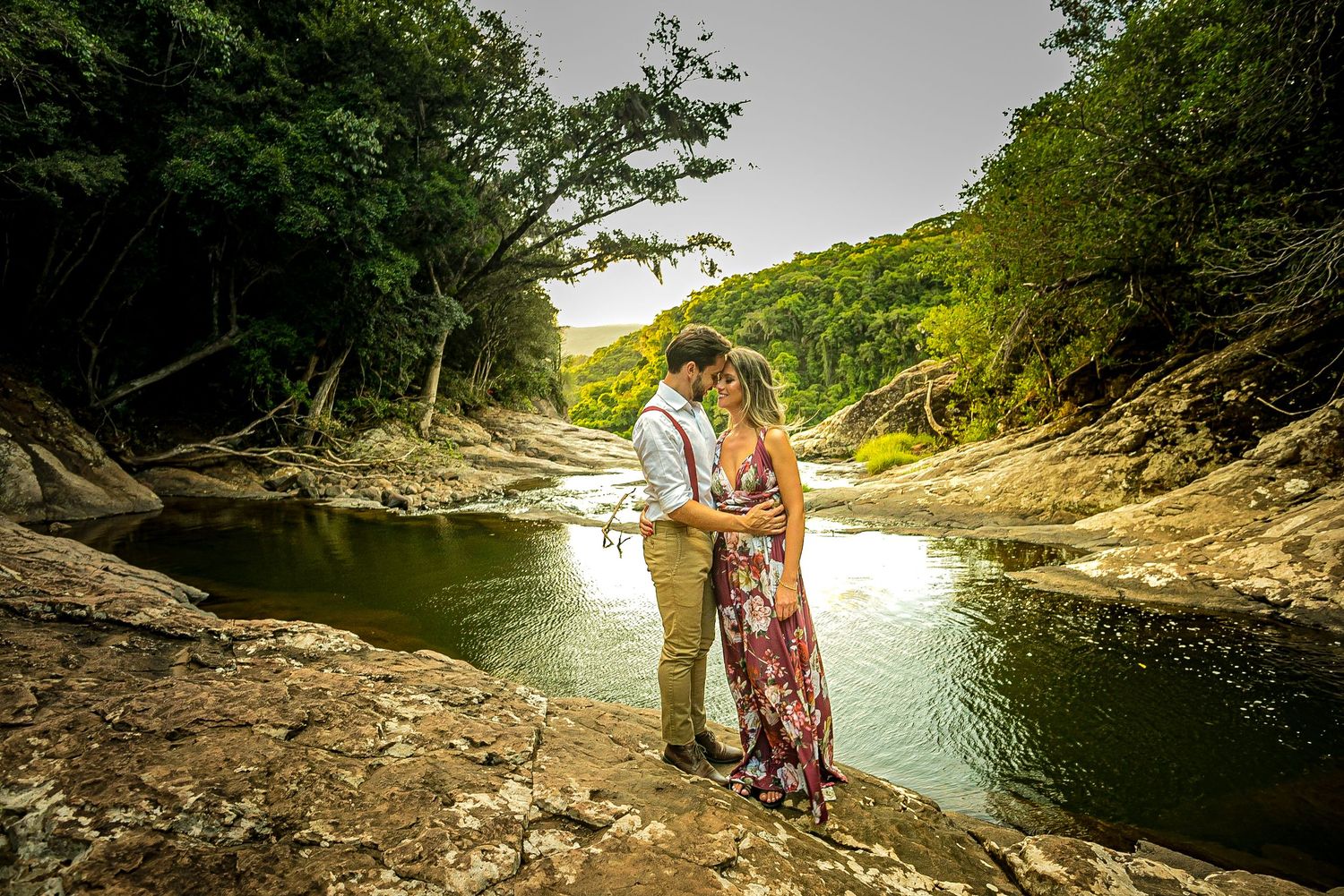 A couple sharing an intimate moment on rocky riverbank surrounded by forest.