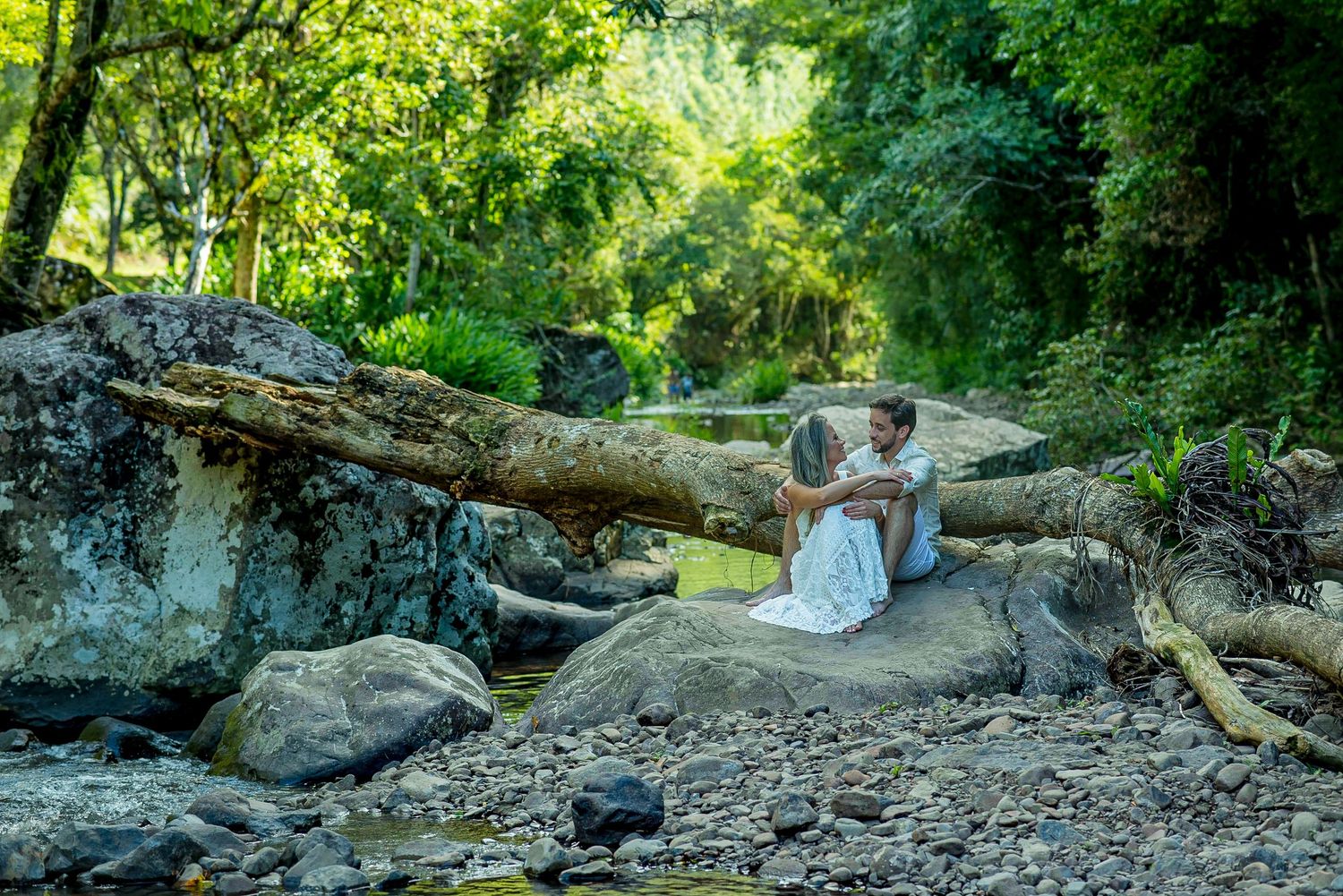 A couple sitting on a large log by a rocky creek bed in a green forest setting.