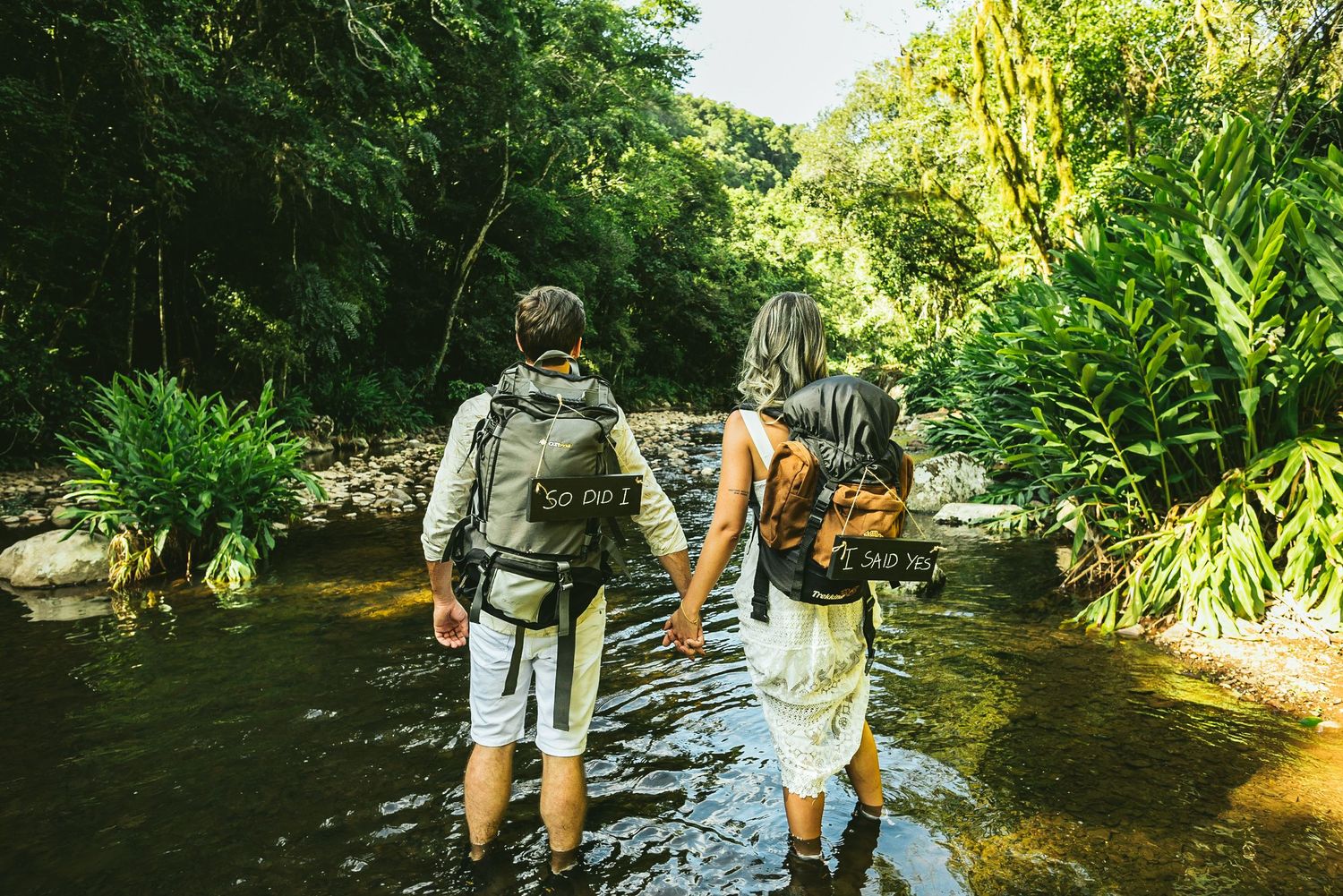 Adventurous couple hiking through a stream with backpacks in a lush forest.