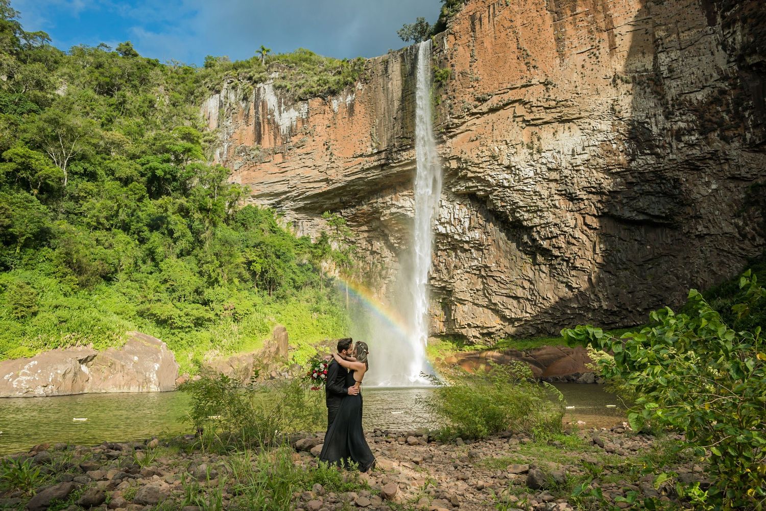 A beautiful rainbow appears beside a towering waterfall as two figures stand at its base.