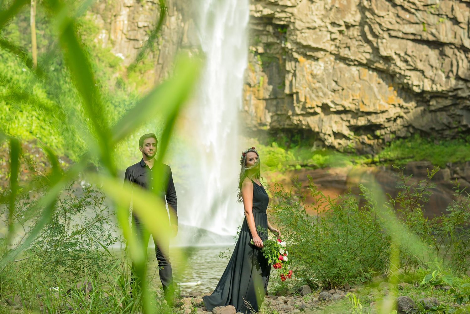 Two people pose near dramatic waterfall with lush greenery in foreground.