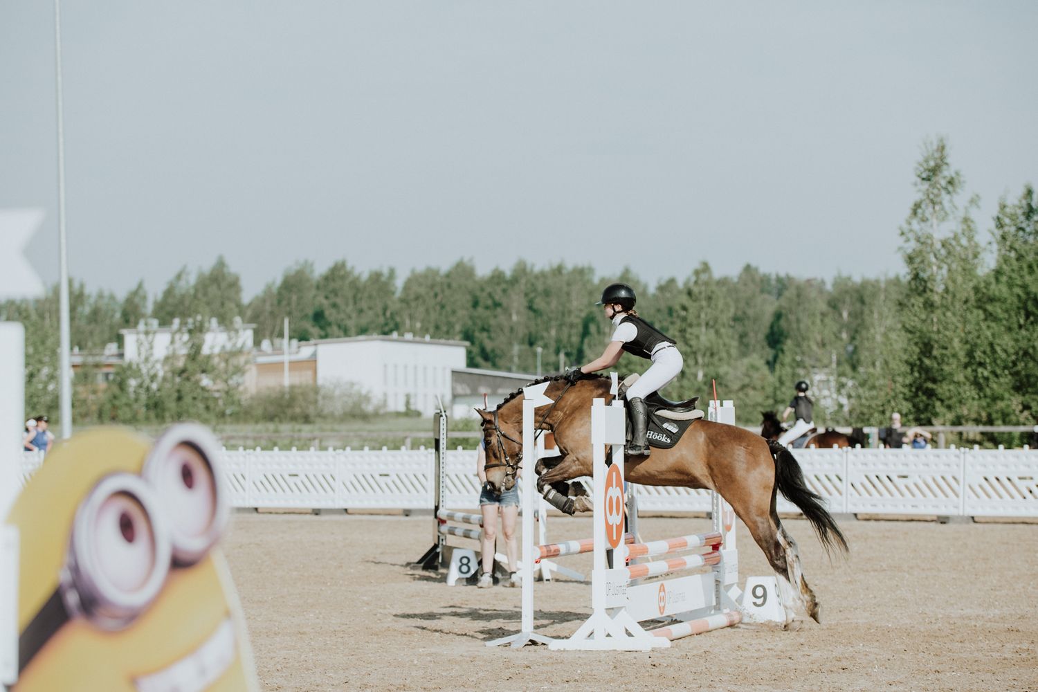 Horse and rider clearing a show jumping fence with Minion-themed course decorations.