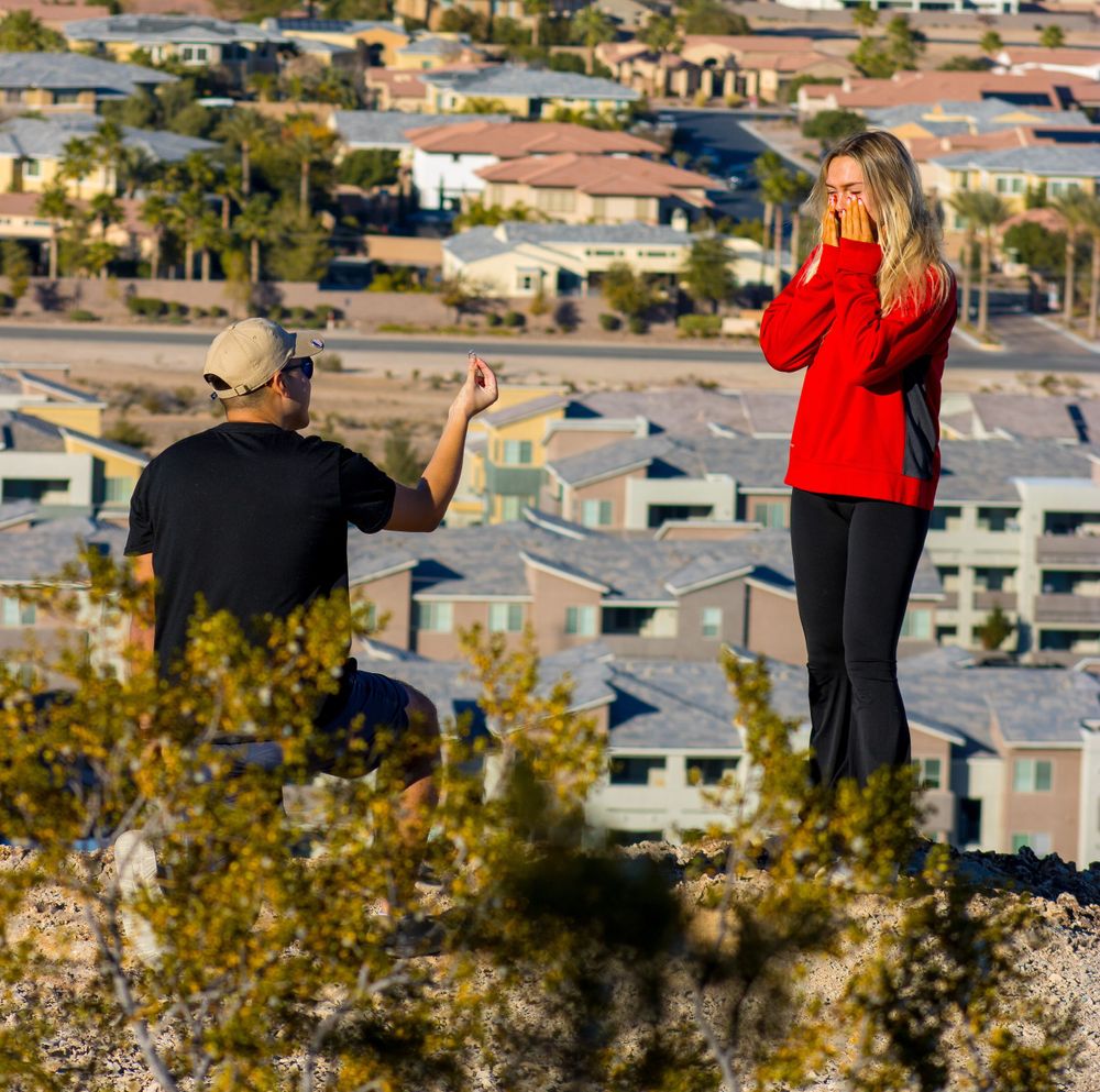 Man kneeling to propose to surprised woman on a rocky hilltop overlooking a suburban Las Vegas neighborhood.