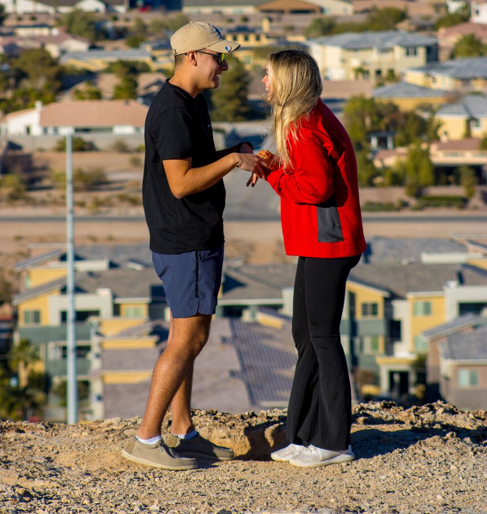 Man placing engagement ring on womans finger during a romantic proposal on a scenic desert hilltop.