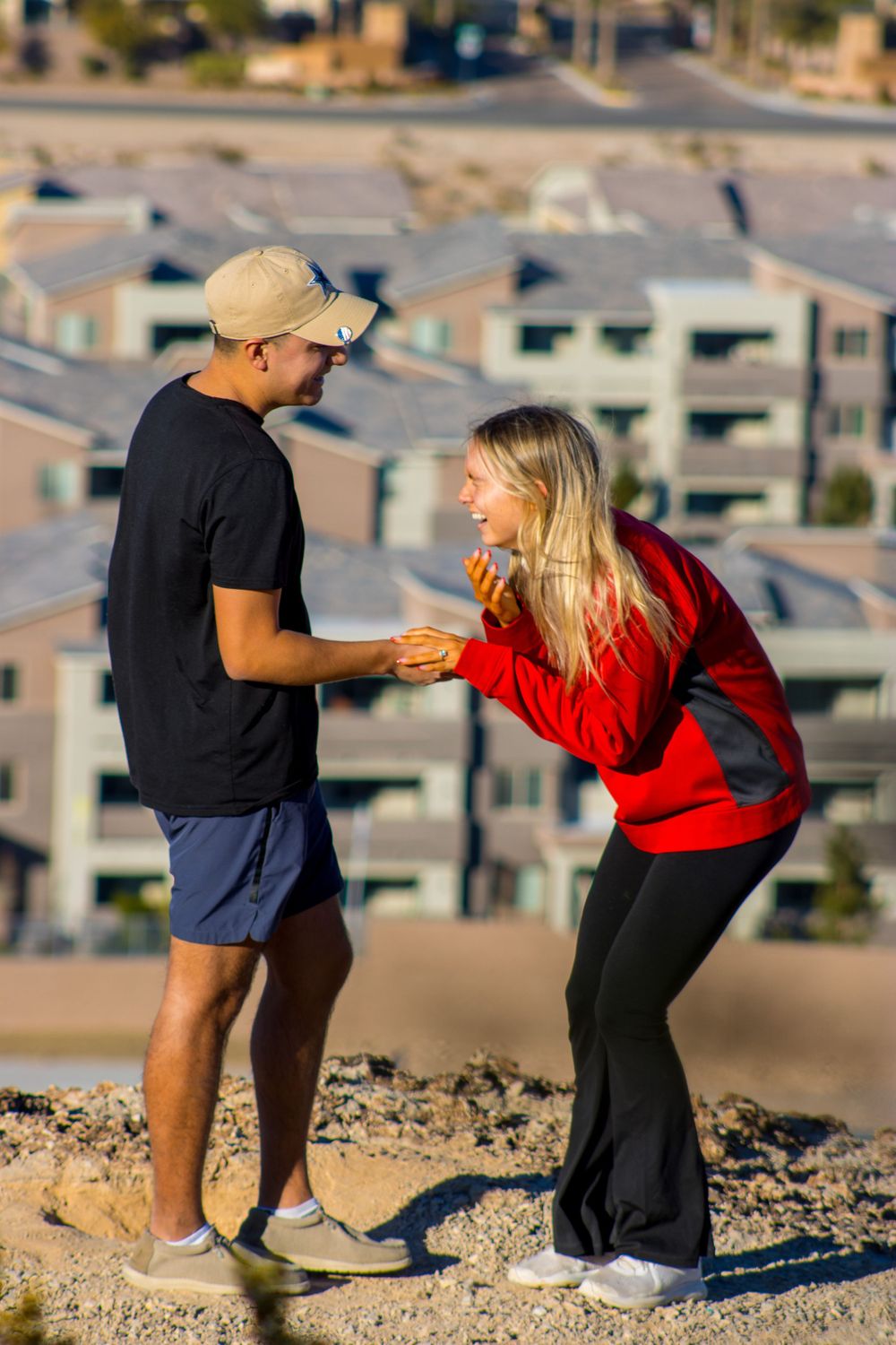 Couple laughing joyfully together after an engagement proposal on a rocky overlook above a city neighborhood.