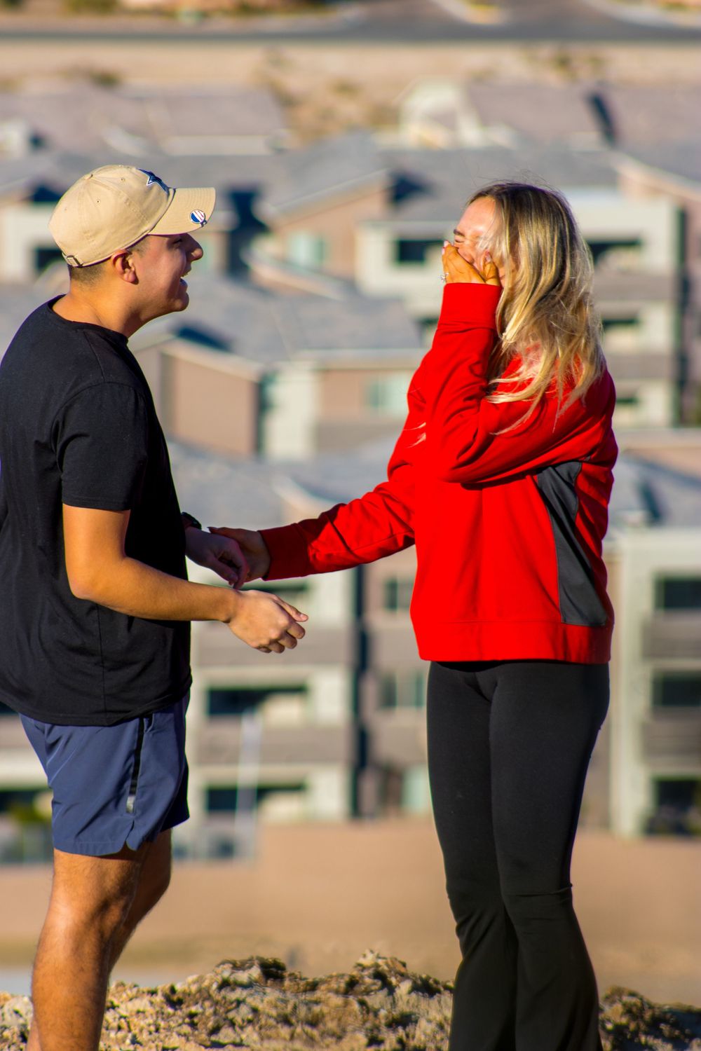 Newly engaged couple holding hands and smiling at each other on a hilltop after a surprise proposal.