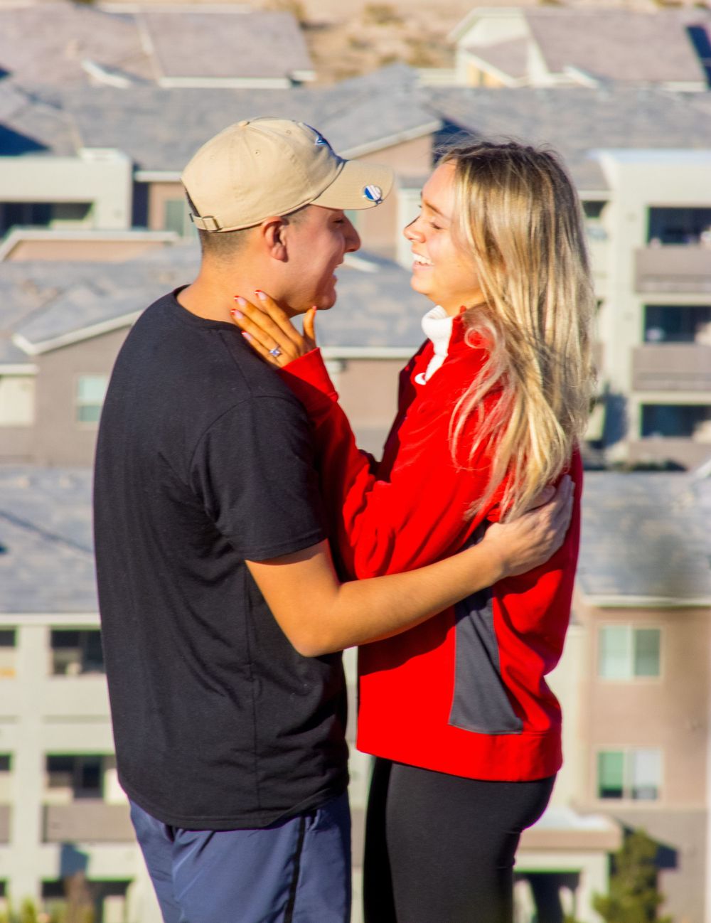 Young couple in red and black outfits sharing romantic moment outdoors with residential buildings behind.