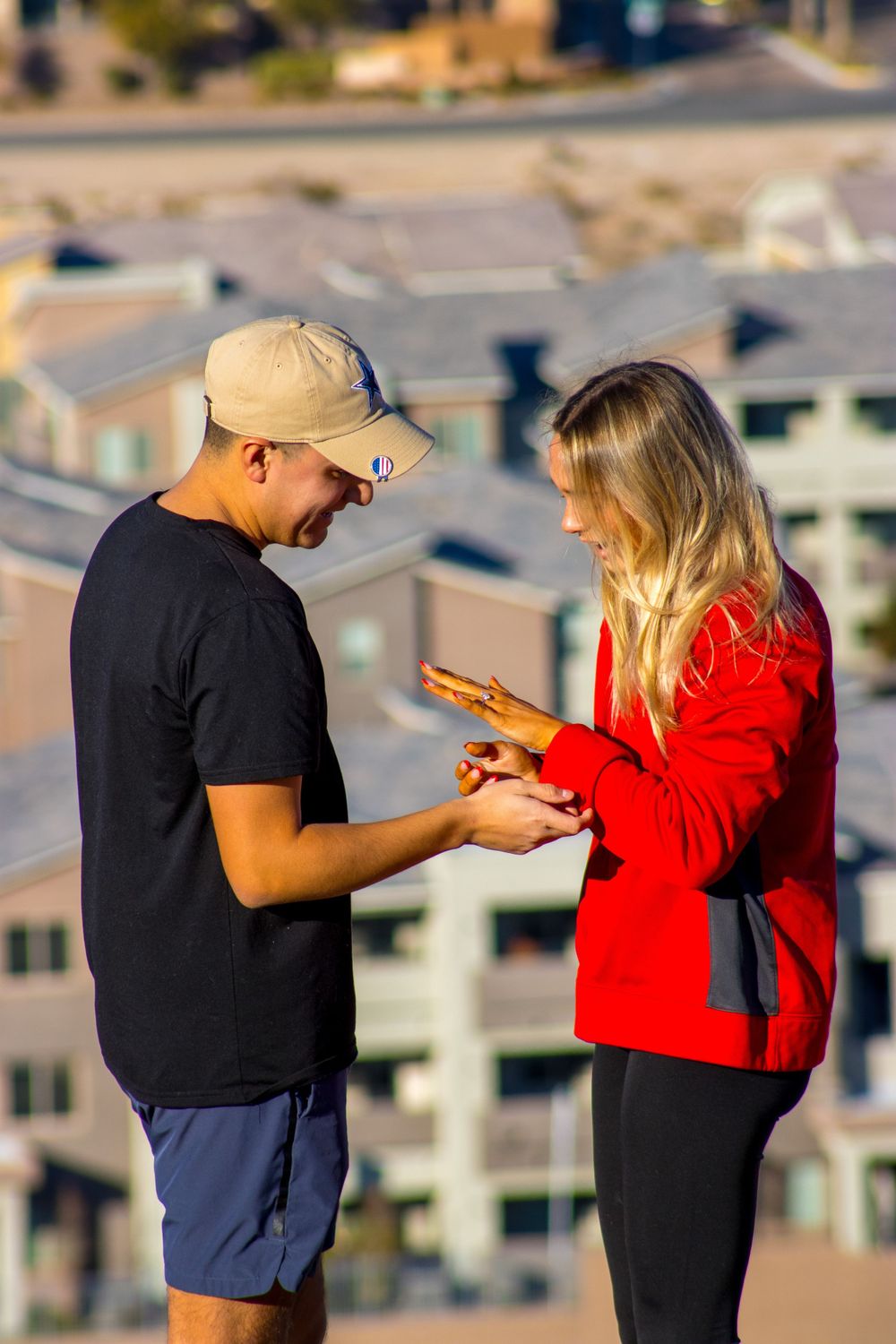 Man placing engagement ring on woman's finger during outdoor proposal with buildings in background.