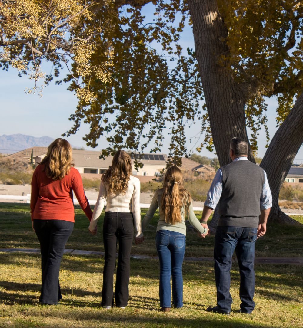 Family of four holding hands while standing under a tree, viewed from behind in autumn park.