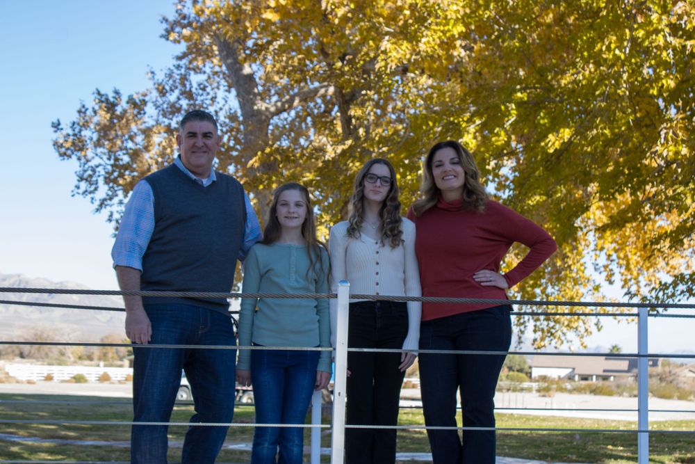 Family of four smiling together by a white fence with golden autumn trees in background.